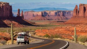 A white SUV with gear on its roof journeys down a meandering road through the Desert Southwest, flanked by red rock formations, mesas, and tall cacti. The sky is partly cloudy, and distant mountains rise in the background, evoking the spirit of a classic road trip adventure.